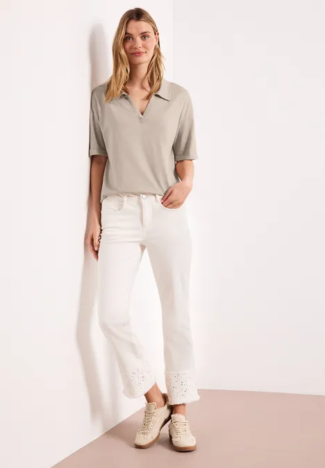 Frau lehnt an weißer Wand, trägt graues Poloshirt, weiße Jeans mit Spitzensaum und helle Sneakers.