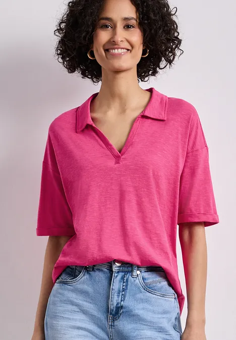 Vrouw met krullend haar in roze poloshirt en lichtblauwe jeans voor een neutrale achtergrond.