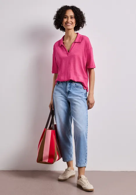 Vrouw in roze blouse, lichtblauwe jeans en sneakers, met een grote tweekleurige tas voor een neutrale achtergrond.