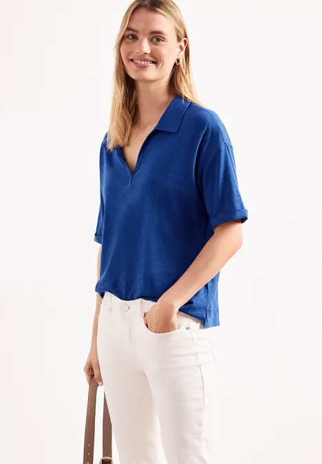 Eine Frau trägt ein blaues Poloshirt mit kurzen Ärmeln, weiße Hose und hält eine beige Tasche vor neutralem Hintergrund.