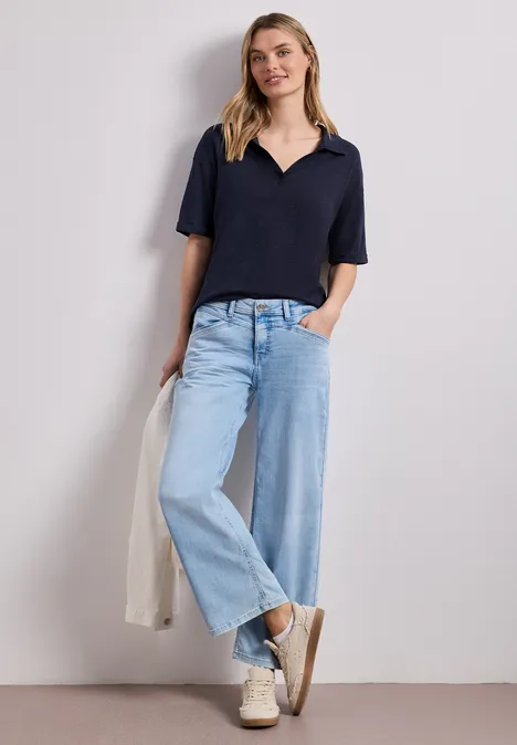 Frau in lässigem Outfit: dunkelblaues Poloshirt, weite Jeans, weiße Sneaker, leichte Jacke in der Hand.