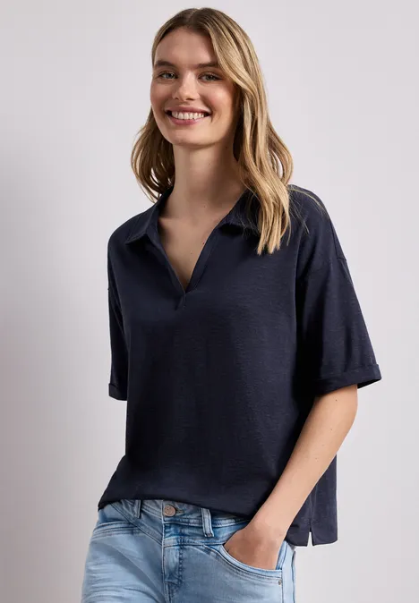 Person vor heller Wand, trägt locker sitzendes dunkelblaues Poloshirt, eine Hand in der Jeans-Tasche.