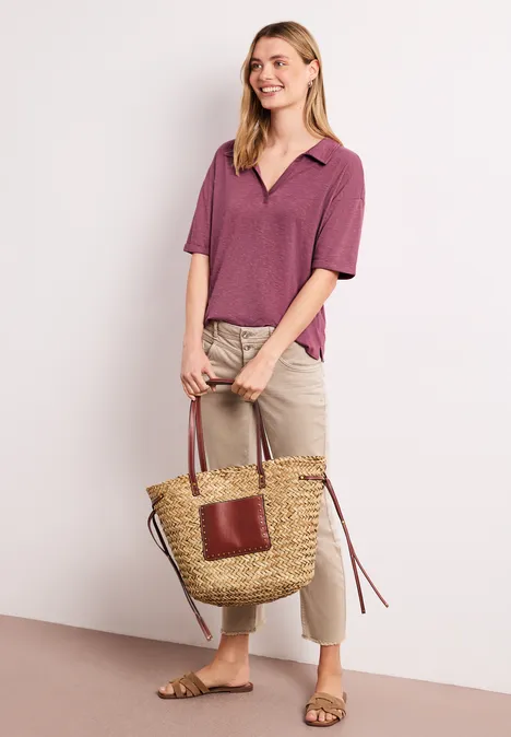 Frau in lila Polo, beigen Hosen und Sandalen trägt geflochtene Tasche vor hellem Hintergrund.