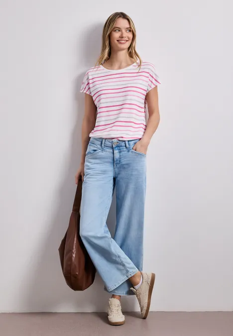 Frau in gestreiftem T-Shirt, weiten Jeans und Sneakern mit brauner Tasche lehnt an weißer Wand.