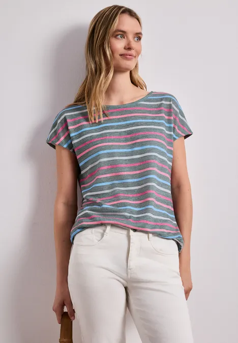 Person trägt bunt gestreiftes T-Shirt und helle Hose vor neutraler Wand.