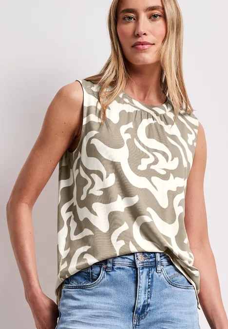 Vrouw in mouwloos beige-wit dessin topje en blauwe spijkerbroek voor een lichte achtergrond.