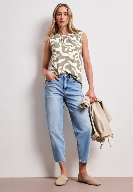 Vrouw in mouwloos top met print, wijde high-waist jeans, beige instappers, houdt een beige jasje vast.