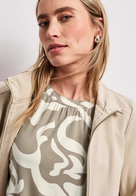 Persoon met lang, blond haar draagt een beige leren jas en een olijfgroen shirt met wit dessin.