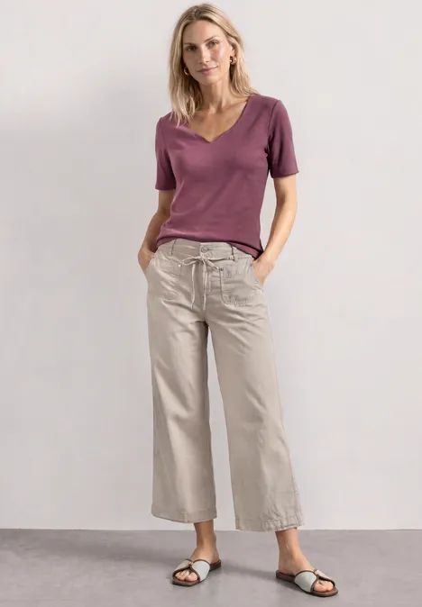 Basic T-Shirt mit Herzausschnitt dark clouded blush Frau in lila T-Shirt und hellen, weiten Hosen steht vor neutralem Hintergrund.