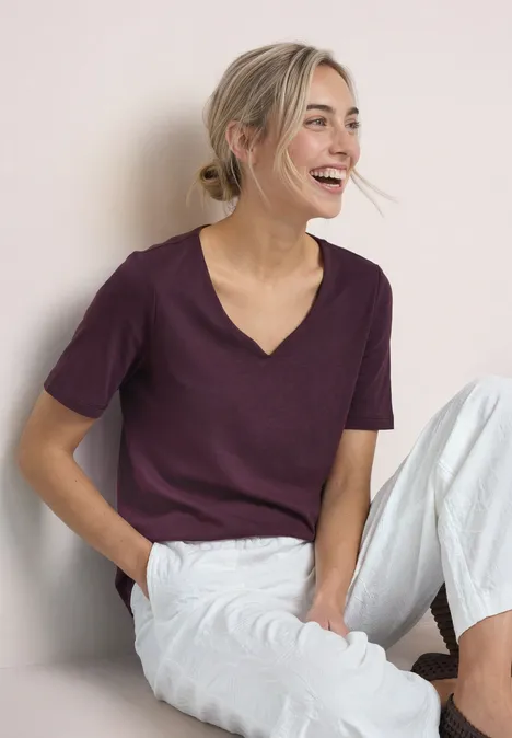 Basic T-Shirt mit Herzausschnitt rustic rouge Lächelnde Frau sitzt entspannt in weißer Hose und violettem T-Shirt.