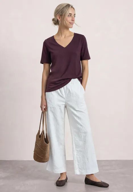 Basic T-Shirt mit Herzausschnitt rustic rouge Frau in lockerem Outfit mit dunkelrotem T-Shirt, weiten weißen Hosen, flachen Schuhen und geflochtener Tasche.