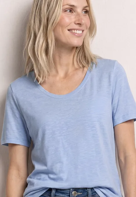 Frau trägt hellblaues T-Shirt und Jeans, zu sehen von Schultern bis Hüfte, vor hellem Hintergrund.