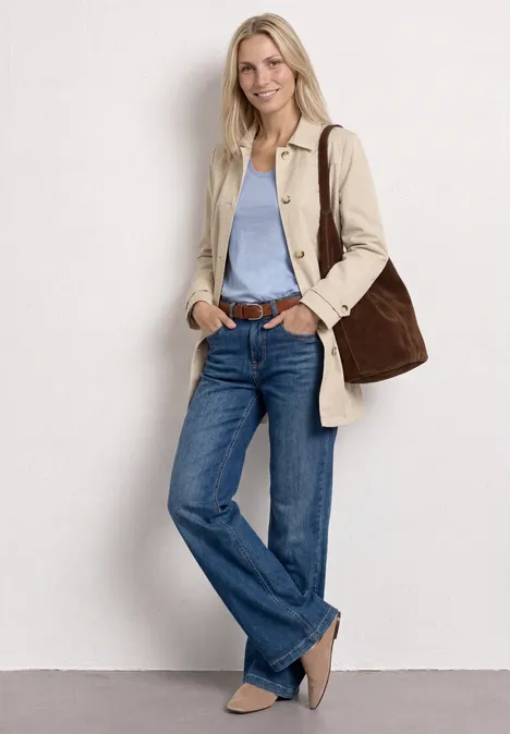 Frau lehnt lässig an weißer Wand, trägt beigen Trenchcoat, hellblaues Shirt, Jeans, beige Ballerinas und große Tasche.