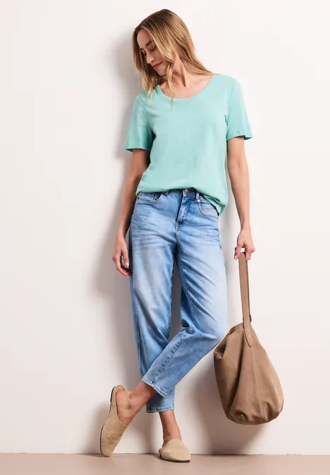 Frau lehnt an Wand mit mintfarbenem Shirt, weiten Jeans, beigen Espadrilles und großer Tasche.