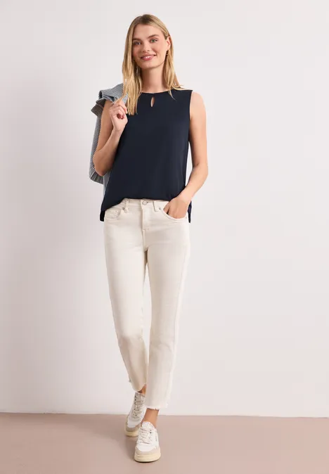Een vrouw in een donkerblauw mouwloos topje, lichte broek en witte sneakers, met tas over de schouder.