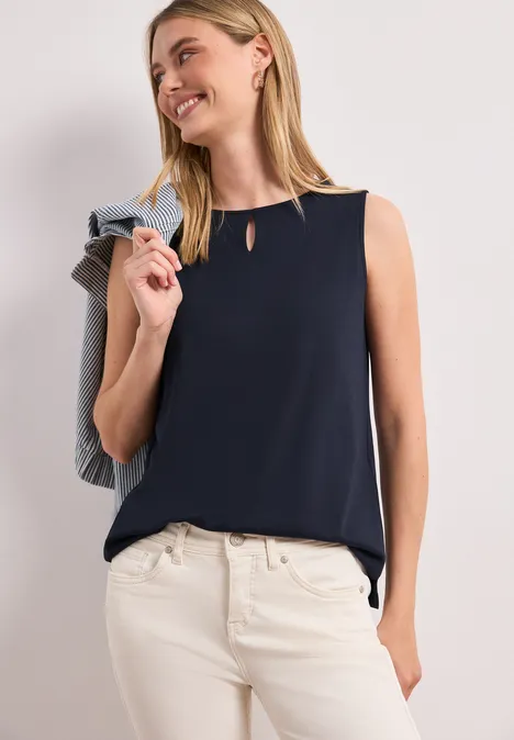 Vrouw in mouwloze donkerblauwe top en witte broek, nonchalant een jasje over de schouder gehangen.
