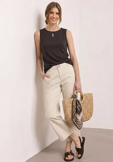 Femme en top noir et pantalon beige, sandales noires, tenant un sac en paille avec foulard.