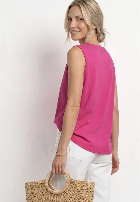 Persoon met roze mouwloos topje, witte broek en ronde rieten tas voor een lichte achtergrond.