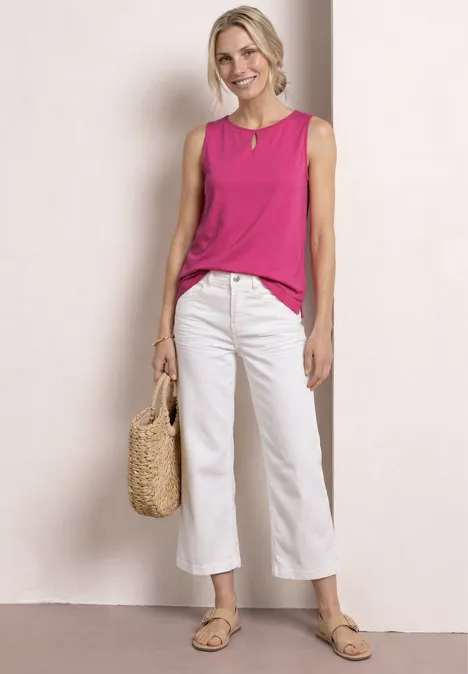 Vrouw met roze mouwloze top, witte culotte, strooien tas en sandalen voor een lichte achtergrond.