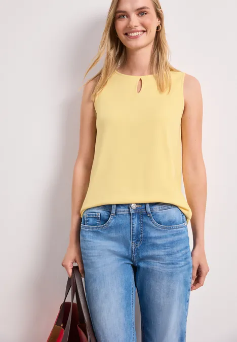 Vrouw met gele mouwloze top en blauwe jeans, draagt een handtas.