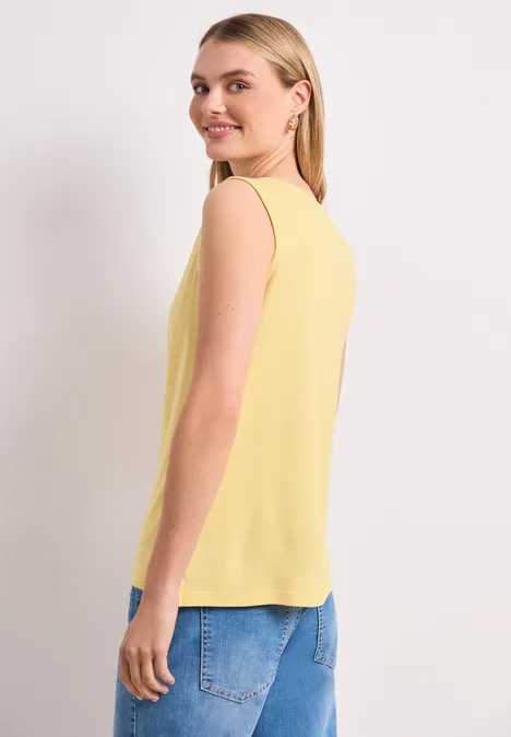 Een vrouw draagt een geel mouwloos topje en een blauwe spijkerbroek, zijwaarts poseerend voor een witte achtergrond.