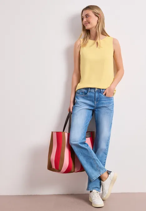 Vrouw in jeans, geel top en witte sneakers leunt tegen muur, draagt gestreepte tas.