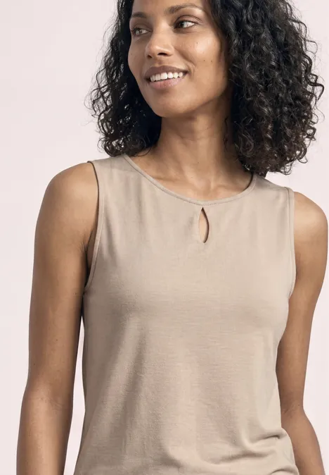 Eine Frau mit lockigen Haaren trägt ein beiges, ärmelloses Top mit kleinem Schlitz am Ausschnitt.