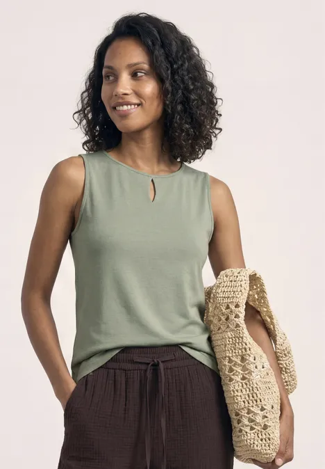 Vrouw met krullend haar draagt een groen mouwloos topje, een bruine broek en een gehaakte tas.