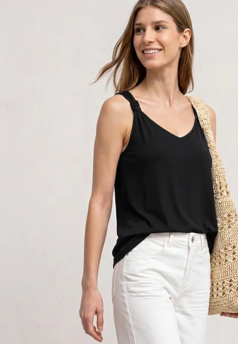 Vrouw in zwarte mouwloze top, witte broek en strooien tas over de schouder.