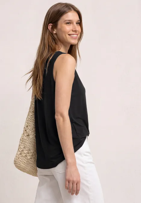 Vrouw in zijaanzicht met zwarte mouwloze top, witte broek en een gevlochten tas van natuurlijk materiaal.