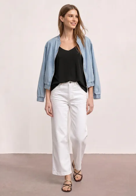 Een vrouw in een zwart topje, witte broek, open spijkerjack en sandalen tegen een lichte muur.