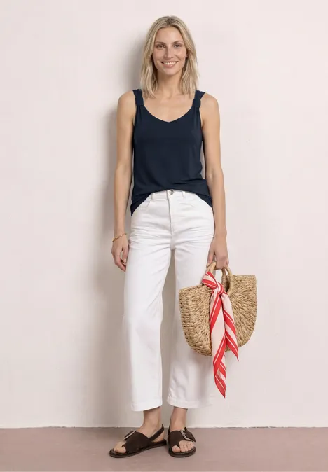 Top mit V-Neck und Knotendetail deep blue Frau in ärmellosem Top, weißen Hosen, schwarzen Sandalen, mit Strohtasche und rotem Tuch steht vor hellem Hintergrund.