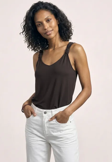 Vrouw met krullend haar draagt een bruine tanktop en witte jeans, handen in de zakken.