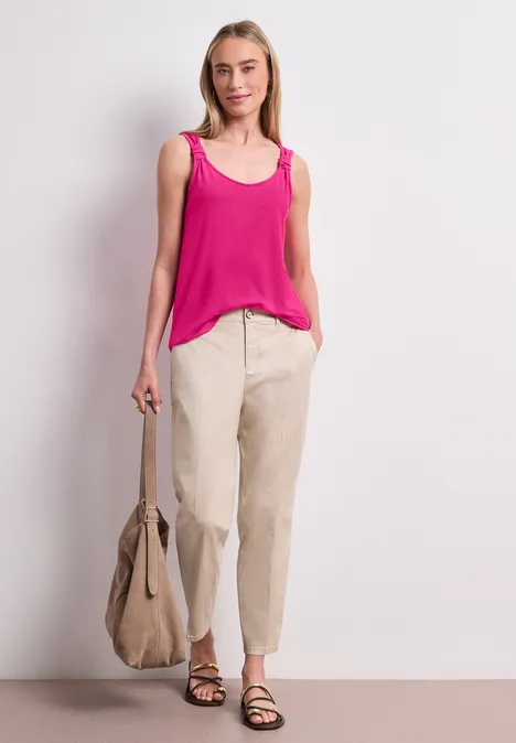 Vrouw in roze hemdje, beige broek en sandalen, met beige tas in hand, staand voor een witte achtergrond.