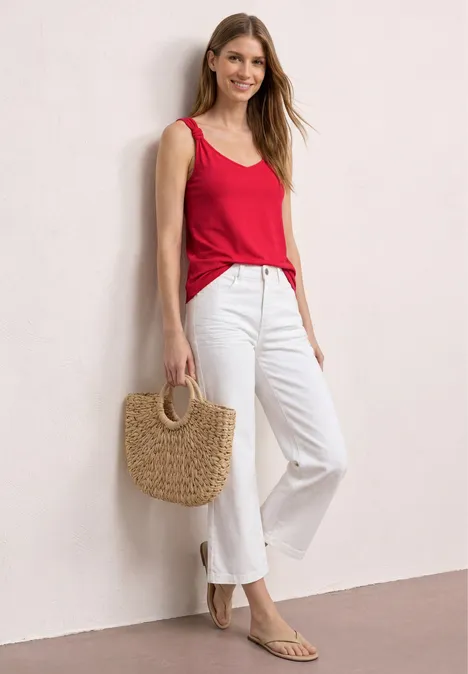 Top mit V-Neck und Knotendetail salsa red Frau lehnt lässig an Wand, trägt rotes Top, weiße Hose, Strohtasche und flache Sandalen.