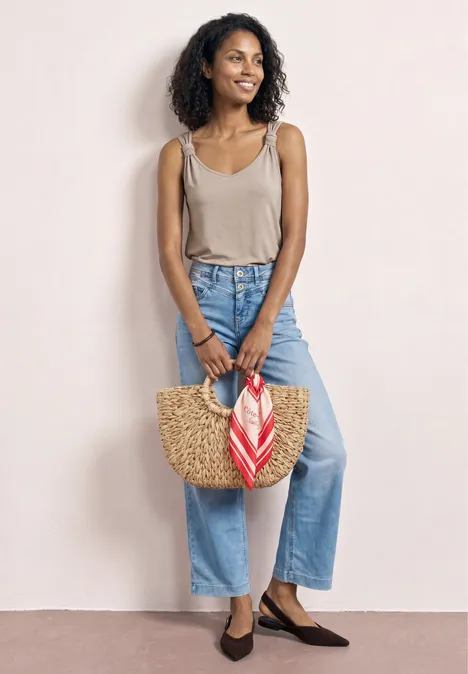 Top mit V-Neck und Knotendetail clay sand Eine Frau in beige Tanktop und hellblauen Jeans hält eine Strohtasche mit rot-weißem Tuch, trägt braune Ballerinas.