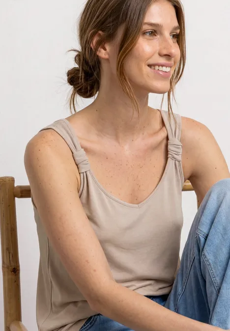 Vrouw zit ontspannen op een stoel, draagt een beige top met knoopschouderbanden en een blauwe jeans.