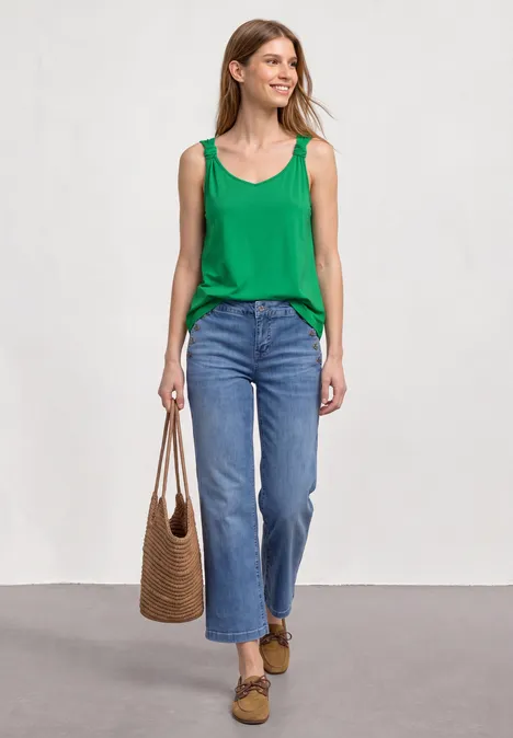 Vrouw in groen topje en blauwe jeans, met rieten tas en bruine loafers voor lichte achtergrond.