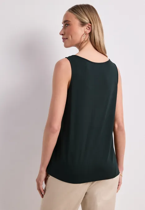 Een vrouw van achteren in een mouwloze donkergroene top en beige broek tegen een neutrale achtergrond.