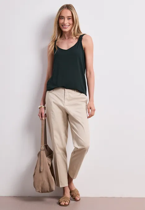 Vrouw in zwart hemdje en beige broek met bijpassende tas voor een witte muur.
