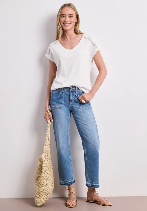 Frau mit weißem T-Shirt, blauen Jeans, flachen Sandalen und Korbtasche, Gesicht unscharf.