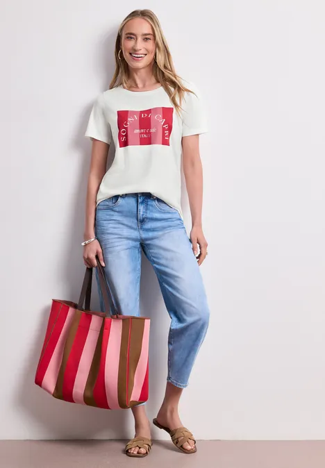 T-Shirt mit Rundhals und Print off white Frau trägt weißes T-Shirt mit rotem Print, hellblaue Jeans, goldene Sandalen und rot-rosa gestreifte Tasche.