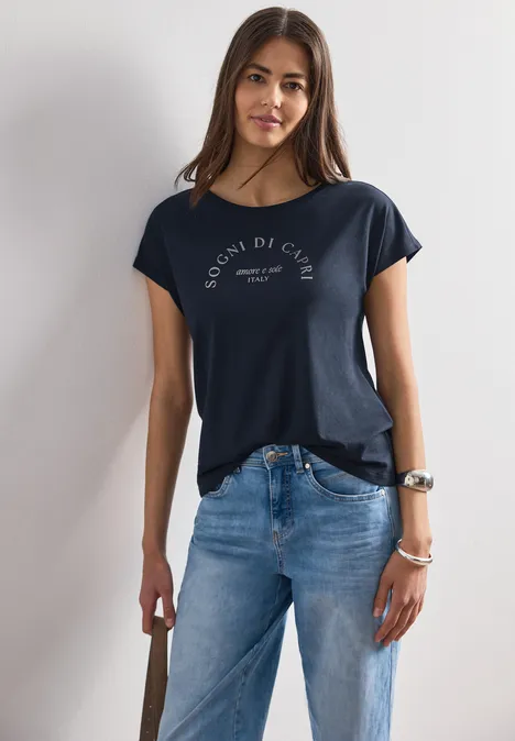Dropped Shoulder Shirt mit Rundhals deep blue Frau in dunklem T-Shirt und blauer Jeans lehnt an weißer Wand.
