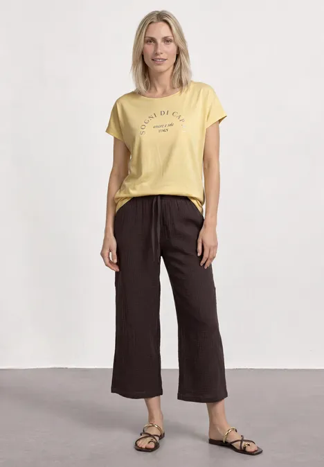 Koszulka na ramiączkach z okrągłym dekoltem soft yellow Kobieta w żółtej koszulce, ciemnych szerokich spodniach 7/8 i sandałach na jasnym tle.