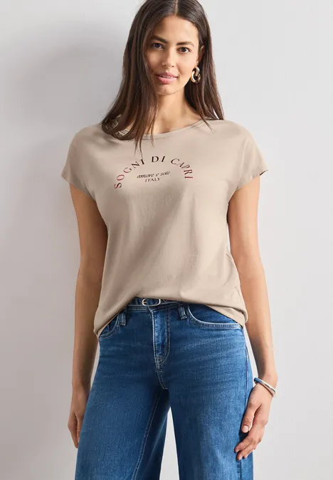 Dropped Shoulder Shirt mit Rundhals clay sand Frau im beigen T-Shirt mit Schriftzug und blauen Jeans vor hellem Hintergrund.