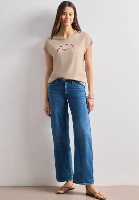 Dropped Shoulder Shirt mit Rundhals clay sand Frau in beigem T-Shirt, weiten blauen Jeans und Sandalen, vor weißem Hintergrund.