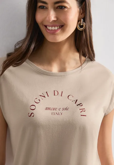 Dropped Shoulder Shirt mit Rundhals clay sand Fröhliche Frau mit hellbeigem T-Shirt mit Aufdruck 'SOGNI DI CAPRI amore e sole ITALY' und goldenen Creolen