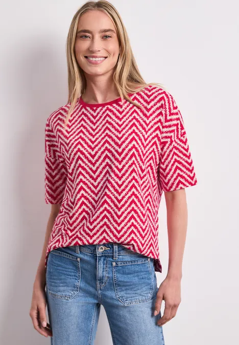 Frau im rot-weißem Zickzack-Shirt und blauen Jeans vor hellem Hintergrund.