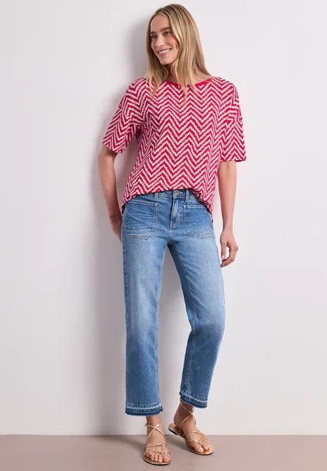Frau in rotem Zickzack-T-Shirt, blauen Cropped-Jeans und goldenen Sandalen vor neutralem Hintergrund.