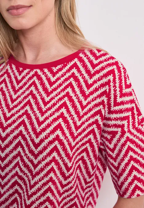 Frau trägt rotes T-Shirt mit weißem Zickzackmuster, halblange Ärmel und Rundhalsausschnitt.